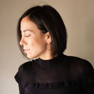 Puede incluir: Una persona con un jersey negro de cuello alto y grandes pendientes de aro plateados. Los pendientes son circulares. La persona tiene el pelo corto y oscuro y mira hacia abajo. El fondo es una pared lisa.