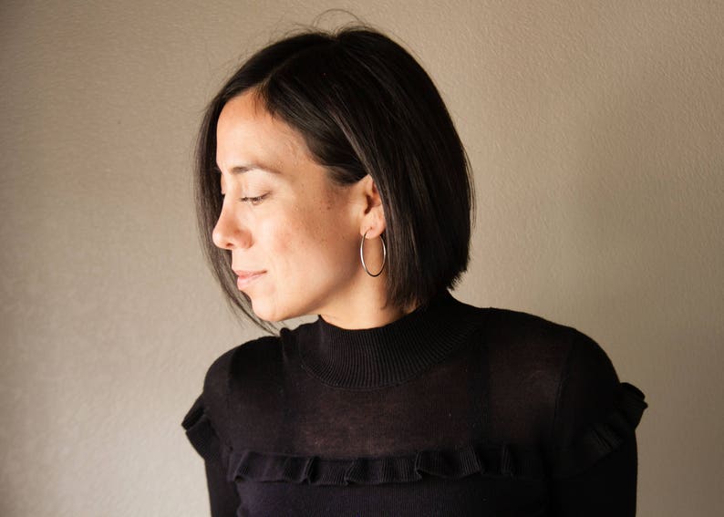 Puede incluir: Una persona con un jersey negro de cuello alto y grandes pendientes de aro plateados. Los pendientes son circulares. La persona tiene el pelo corto y oscuro y mira hacia abajo. El fondo es una pared lisa.