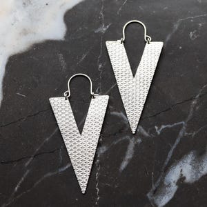 May include: A pair of silver earrings in a V-shape design. The earrings have a textured surface and hang from a simple hook. The background is a dark, marbled surface. The earrings are made of metal.