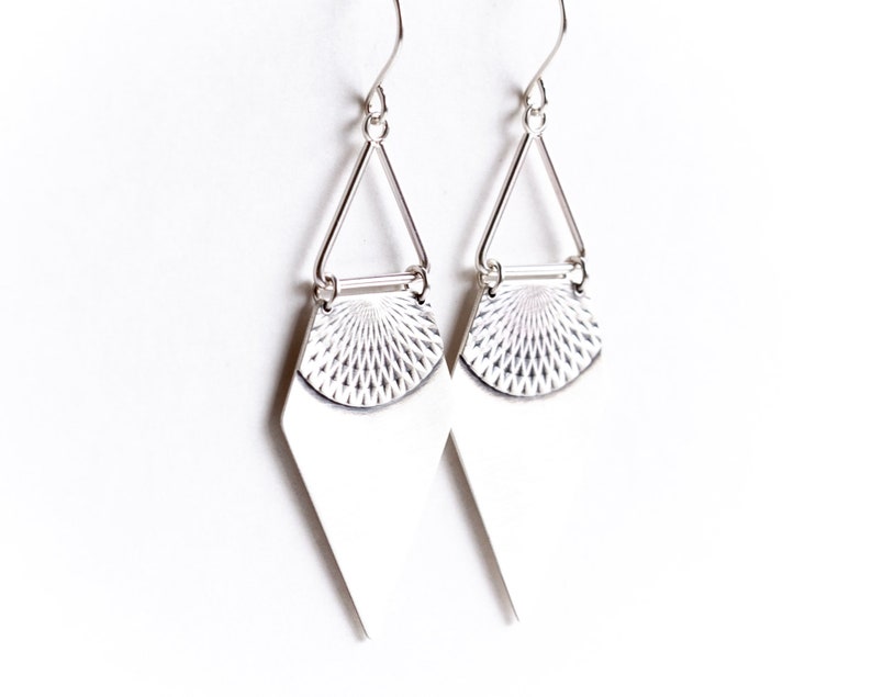May include: A pair of silver earrings with a geometric design. Each earring features a diamond-shaped bottom, a textured half-circle, and a triangular top connected to a hook. The earrings are displayed against a white background.