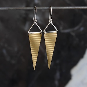 May include: A pair of geometric earrings. Each earring features a silver triangle top with a horizontal bar, connected to a gold-toned, striped, downward-pointing triangle. The earrings hang from silver hooks. The background is dark and blurred.