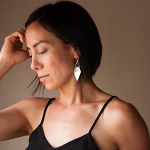 May include: Silver geometric earrings with a diamond shape and a triangular top are displayed. The earrings are hanging from a woman's ear. A silver bracelet is on the woman's wrist. The woman is wearing a black tank top.