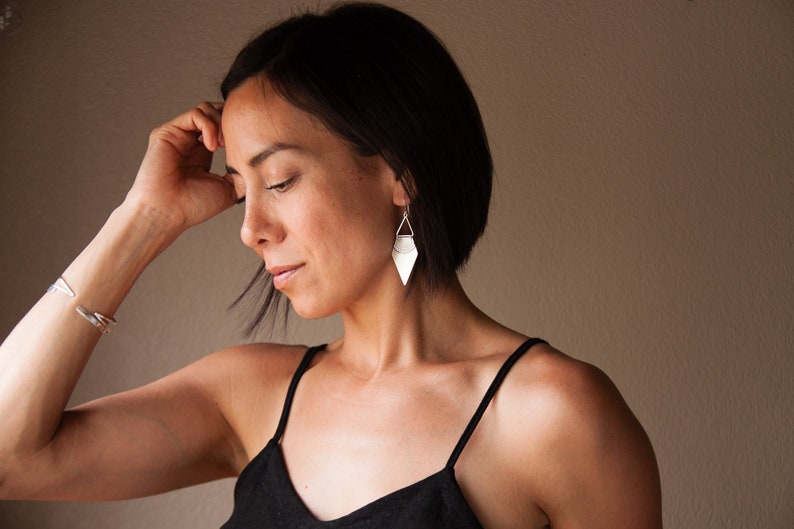 May include: Silver geometric earrings with a diamond shape and a triangular top are displayed. The earrings are hanging from a woman's ear. A silver bracelet is on the woman's wrist. The woman is wearing a black tank top.