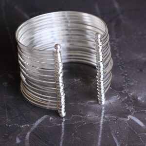 May include: A silver-colored cuff bracelet with multiple parallel bands. The bracelet has a vertical support structure on each side, with a small ball at the top. The jewelry is displayed on a dark, marbled surface.