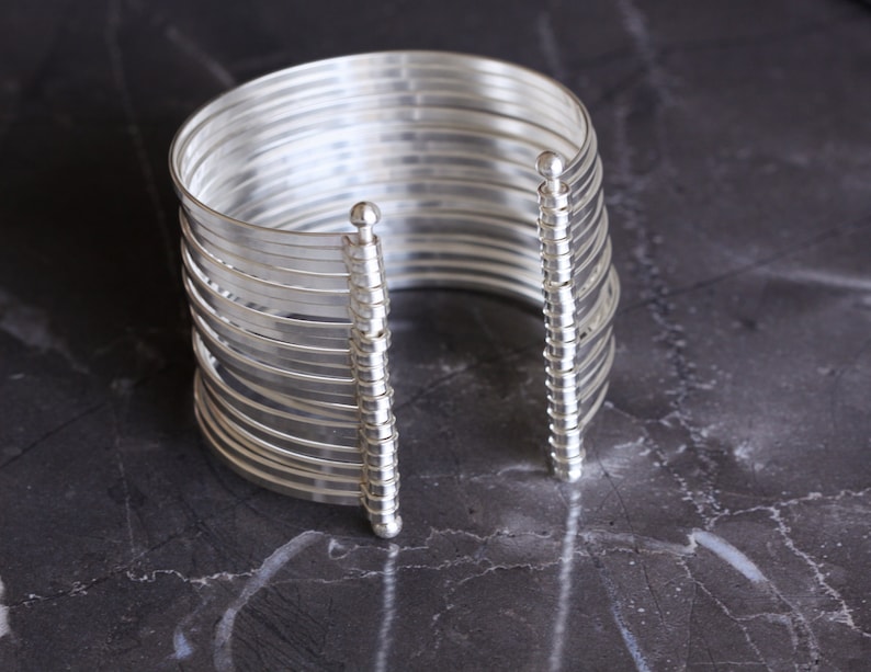 May include: A silver-colored cuff bracelet with multiple parallel bands. The bracelet has a vertical support structure on each side, with a small ball at the top. The jewelry is displayed on a dark, marbled surface.