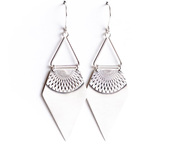 May include: A pair of silver-toned earrings. Each earring features a geometric design with a triangular top, a textured semi-circle, and a diamond-shaped bottom. The earrings hang from a simple hook.