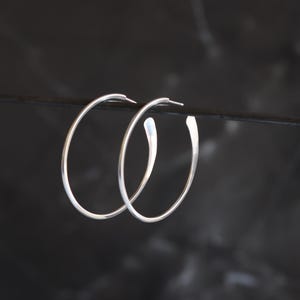 Puede incluir: Un par de pendientes de aro plateados. Los pendientes tienen un dise&ntilde;o minimalista con un acabado liso y pulido. Los aros son redondos y tienen una ligera curva en la parte inferior. Los pendientes se muestran sobre un fondo oscuro.