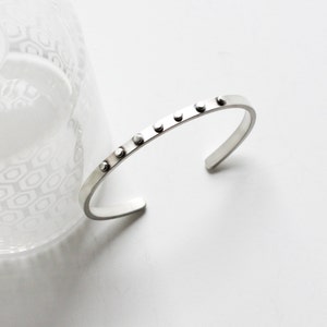 May include: A silver cuff bracelet with a row of small, evenly spaced, circular studs. The bracelet has a simple, minimalist design and is open at the bottom. The background is white with a blurred glass object.