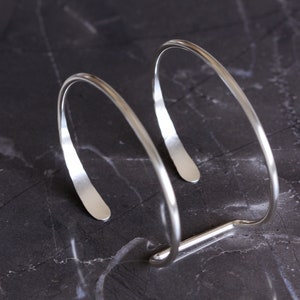 Puede incluir: Una pulsera de pu&ntilde;o plateada con un dise&ntilde;o minimalista. La pulsera tiene un dise&ntilde;o curvo y abierto con un extremo plano y rectangular en cada lado. La pulsera mide aproximadamente 6,35 cm de ancho.