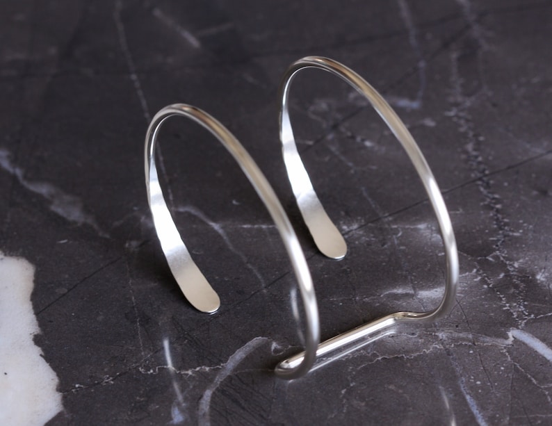 Puede incluir: Una pulsera de pu&ntilde;o plateada con un dise&ntilde;o minimalista. La pulsera tiene un dise&ntilde;o curvo y abierto con un extremo plano y rectangular en cada lado. La pulsera mide aproximadamente 6,35 cm de ancho.
