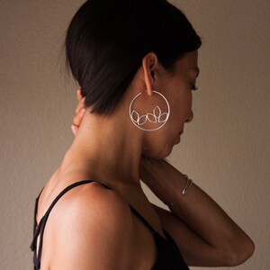 May include: Silver hoop earrings with a delicate leaf design. The earrings are large, circular, and feature a minimalist aesthetic. The leaves are clustered in the lower part of the hoop. The model is wearing a black top.