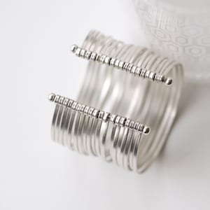 May include: A silver cuff bracelet with multiple parallel bands. The bracelet features two horizontal bars with small, evenly spaced decorative elements and rounded end caps. The jewelry is set against a white background with a soft, patterned fabric in the background.