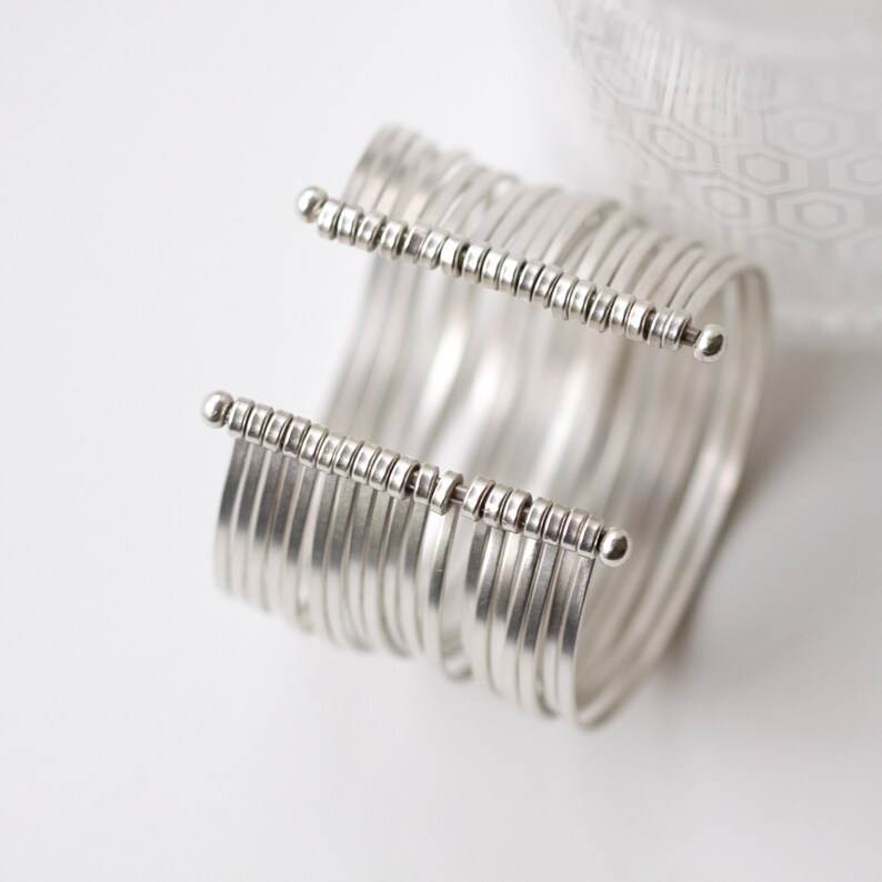 May include: A silver cuff bracelet with multiple parallel bands. The bracelet features two horizontal bars with small, evenly spaced decorative elements and rounded end caps. The jewelry is set against a white background with a soft, patterned fabric in the background.