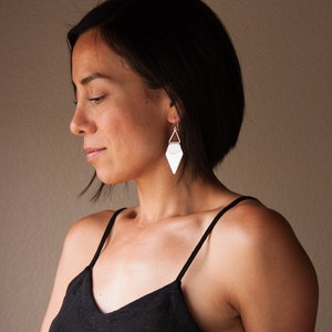 May include: A woman wearing silver geometric earrings. The earrings have a triangular shape with a flat, textured surface and a small triangle at the top. The woman is wearing a black spaghetti strap top.