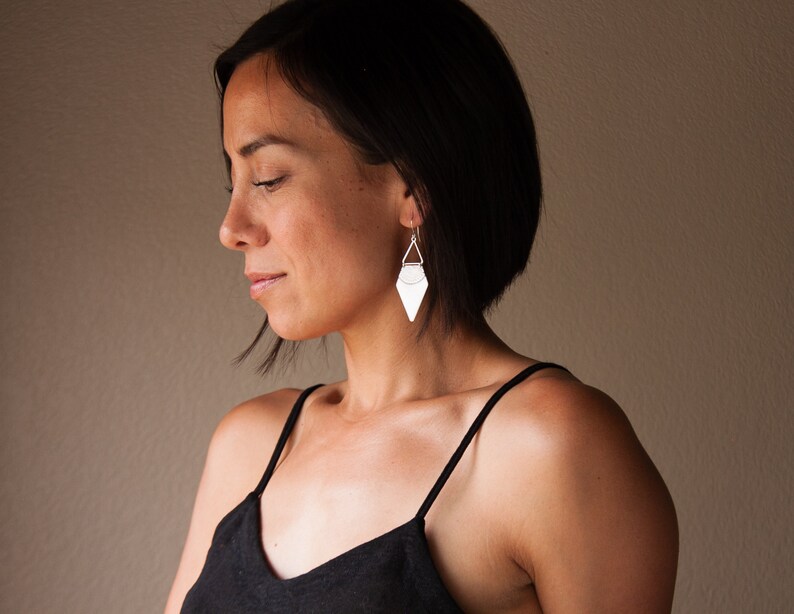 May include: A woman wearing silver geometric earrings. The earrings have a triangular shape with a flat, textured surface and a small triangle at the top. The woman is wearing a black spaghetti strap top.