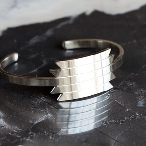 May include: A silver cuff bracelet with a geometric design. The bracelet features a series of horizontal bars that form a chevron shape. The metal has a polished finish, reflecting light. The bracelet is displayed on a dark, textured surface.