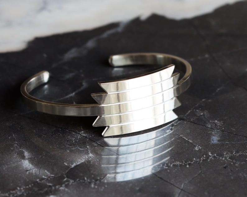 May include: A silver cuff bracelet with a geometric design. The bracelet features a series of horizontal bars that form a chevron shape. The metal has a polished finish, reflecting light. The bracelet is displayed on a dark, textured surface.