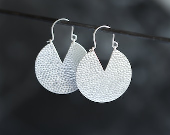 May include: A pair of silver earrings with a hammered texture. Each earring has a circular shape with a V-shaped cutout at the bottom. The earrings hang from silver hooks, and the background is dark.