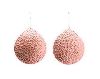 May include: A pair of rose gold teardrop earrings with a textured, hammered metal finish. The earrings hang from silver-colored ear wires. The earrings are displayed against a white background.