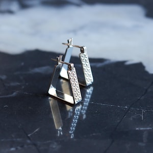 May include: A pair of silver-toned triangle hoop earrings. One earring has a geometric pattern on the vertical bar. The earrings are displayed on a dark, reflective surface, creating a mirrored effect.