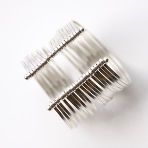 May include: A silver-toned cuff bracelet with a unique design. The bracelet features multiple parallel bands connected by two parallel bars with vertical, thin, silver rods. The design is modern and minimalist.