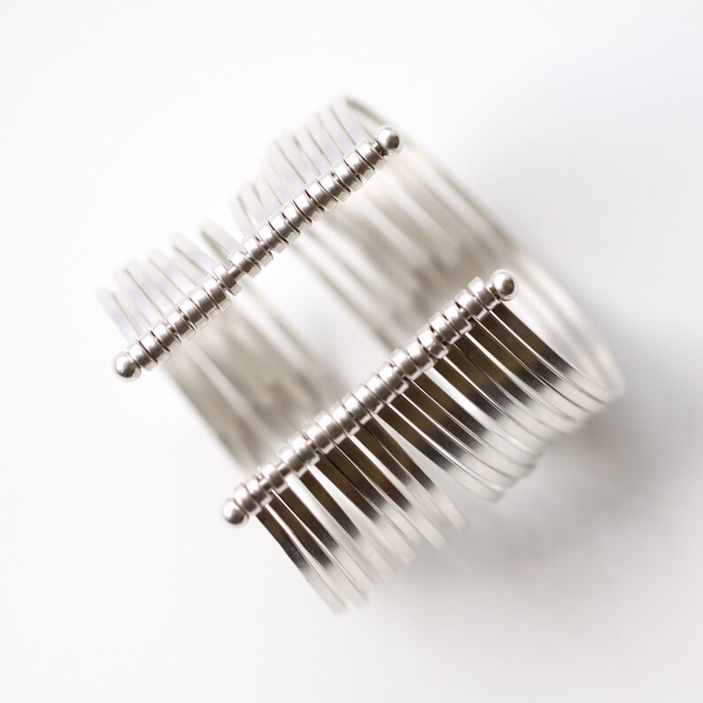 May include: A silver-toned cuff bracelet with a unique design. The bracelet features multiple parallel bands connected by two parallel bars with vertical, thin, silver rods. The design is modern and minimalist.