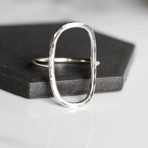 Könnte beinhalten: Ein silberner Ring mit offenem ovalem Design. Der Ring hat eine gehämmerten Textur und liegt auf einer dunkelgrauen Oberfläche. Das Band des Rings ist dünn und verbindet sich mit der ovalen Form. Es ist ein Schmuckstück.