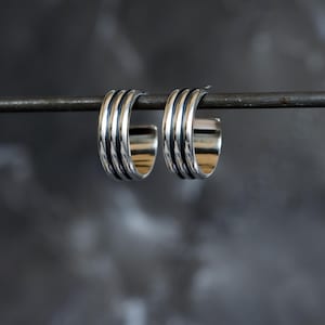 May include: A pair of silver hoop earrings with a ribbed design. Each earring features three parallel, raised bands, creating a textured surface. The earrings are shown against a dark, blurred background, emphasising their metallic shine.
