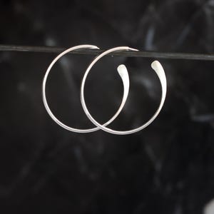 Puede incluir: Un par de pendientes de aro plateados con un dise&ntilde;o en forma de l&aacute;grima. Los pendientes se muestran sobre un fondo oscuro, resaltando su estilo minimalista y elegante. Los aros son finos y tienen un acabado pulido.