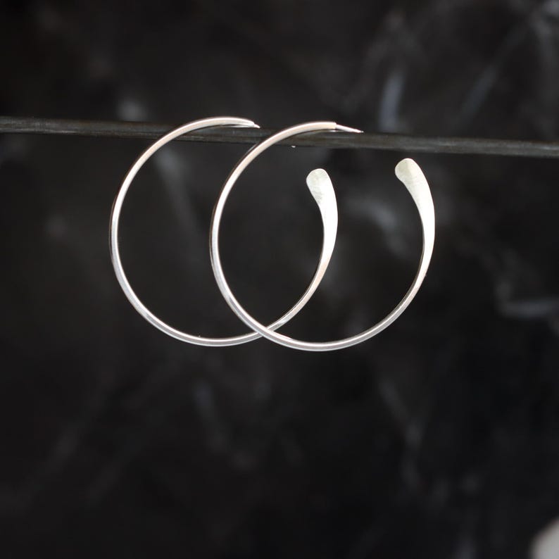 Puede incluir: Un par de pendientes de aro plateados con un dise&ntilde;o en forma de l&aacute;grima. Los pendientes se muestran sobre un fondo oscuro, resaltando su estilo minimalista y elegante. Los aros son finos y tienen un acabado pulido.