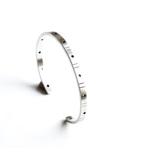 May include: A silver-toned cuff bracelet with a minimalist design. The bracelet features a series of evenly spaced, small circular holes and etched lines along its surface. The open-ended design allows for easy wear. The jewelry is set against a white background.