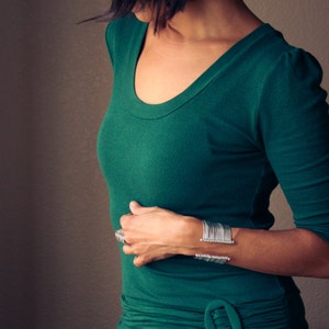 May include: A woman wearing a dark green, scoop-neck top and a silver cuff bracelet. The bracelet has a series of vertical bars. A silver ring is also visible. The top has long sleeves and a fitted silhouette.