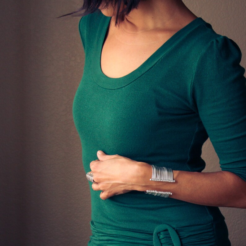 May include: A woman wearing a dark green, scoop-neck top and a silver cuff bracelet. The bracelet has a series of vertical bars. A silver ring is also visible. The top has long sleeves and a fitted silhouette.