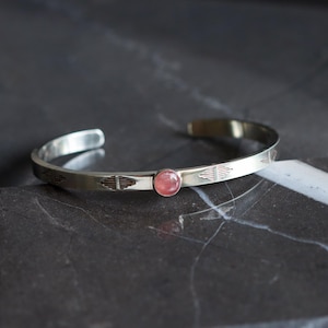 May include: A silver cuff bracelet with a pink gemstone and geometric etched designs. The open-ended bracelet is displayed on a dark surface. The gemstone is a round, light pink color.