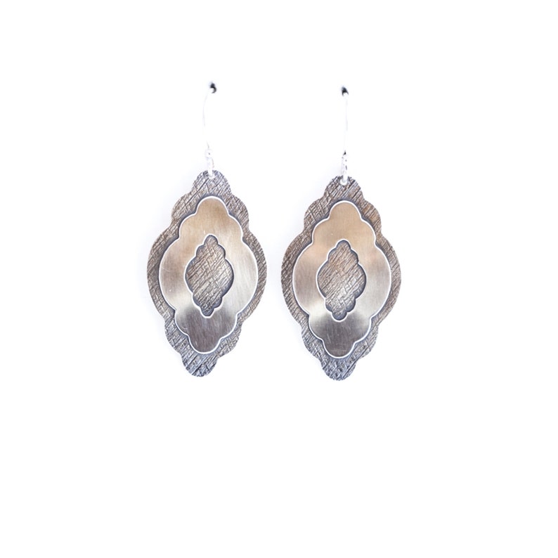 Pode incluir: Um par de brincos de prata com um design em camadas. Cada brinco apresenta uma camada externa texturizada cinza escuro e uma camada interna lisa prateada com uma forma decorativa. Os brincos penduram de ganchos prateados.