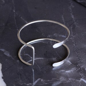 Puede incluir: Dos pulseras de brazalete abiertas de color plateado. Las pulseras est&aacute;n hechas de metal fino y redondeado y tienen un dise&ntilde;o simple y minimalista. Las pulseras descansan sobre una superficie oscura y veteada.