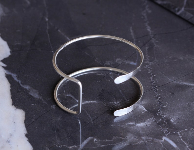 Puede incluir: Dos pulseras de brazalete abiertas de color plateado. Las pulseras est&aacute;n hechas de metal fino y redondeado y tienen un dise&ntilde;o simple y minimalista. Las pulseras descansan sobre una superficie oscura y veteada.