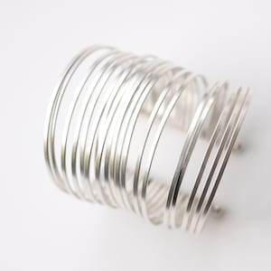 May include: A silver cuff bracelet with multiple parallel bands. The bracelet has a cylindrical shape and is made of shiny metal. The design is modern and minimalist, suitable for various styles. The cuff is shown against a white background.