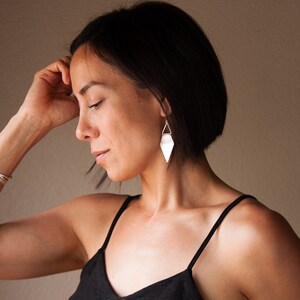 May include: Silver geometric earrings with a triangular shape, hanging from a delicate wire. The earrings are paired with a matching silver bracelet. The woman is wearing a black top.