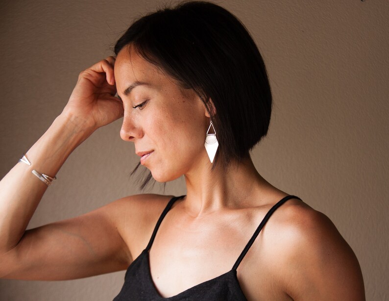 May include: Silver geometric earrings with a triangular shape, hanging from a delicate wire. The earrings are paired with a matching silver bracelet. The woman is wearing a black top.
