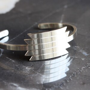 May include: A silver-toned cuff bracelet with a geometric design. The bracelet features a series of layered, angular shapes at its center, creating a bold, modern aesthetic. The metal has a polished finish, reflecting light and adding to its sleek appearance.