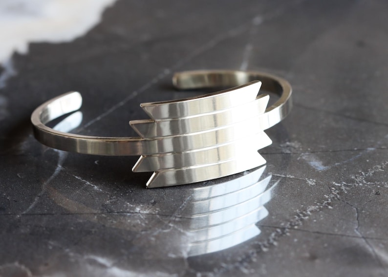 May include: A silver-toned cuff bracelet with a geometric design. The bracelet features a series of layered, angular shapes at its center, creating a bold, modern aesthetic. The metal has a polished finish, reflecting light and adding to its sleek appearance.