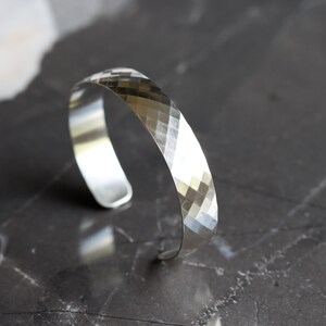 May include: A silver cuff bracelet with a geometric, faceted design. The open-ended bracelet is made of shiny metal and reflects light, creating a sparkling effect. The bracelet is shown on a dark, textured surface.