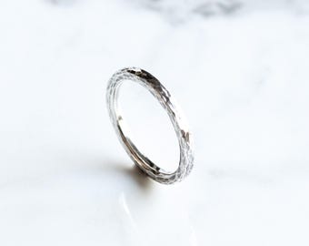 Puede incluir: Primer plano de un anillo de plata con textura martillada. El anillo está sobre una superficie de mármol blanco. La superficie del anillo tiene un patrón texturizado e irregular, que refleja la luz. El anillo es de diseño sencillo.