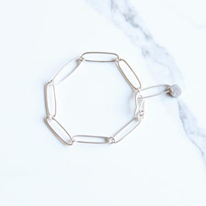 May include: A silver-toned link bracelet with an oval chain design. The bracelet features a small, round charm at the clasp. The jewelry is displayed on a white marble surface.