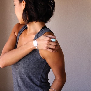 May include: A silver cuff bracelet with a geometric design and a silver ring with a teardrop-shaped, light blue stone. The jewelry is worn on a person's arm and hand, with a gray tank top in the background.