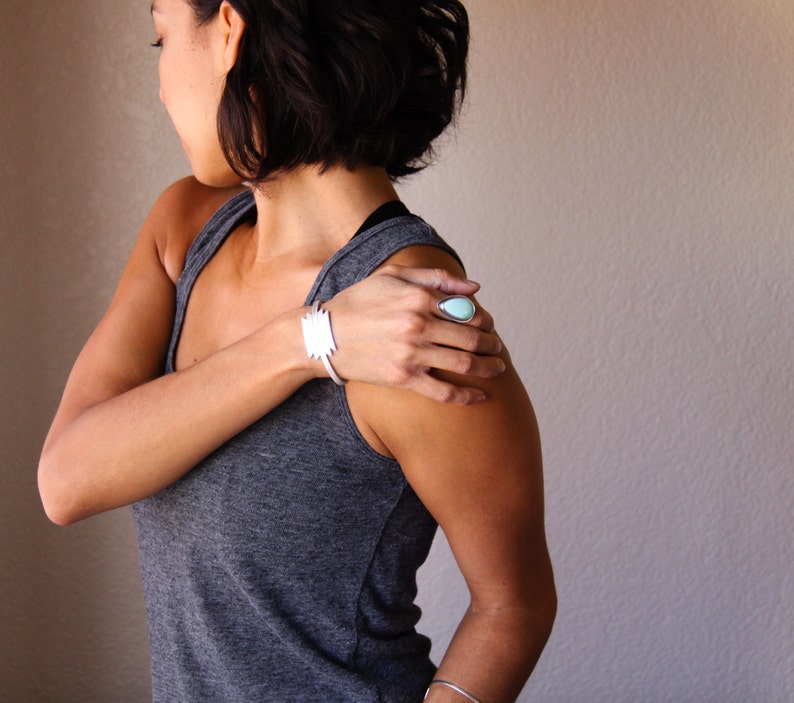 May include: A silver cuff bracelet with a geometric design and a silver ring with a teardrop-shaped, light blue stone. The jewelry is worn on a person's arm and hand, with a gray tank top in the background.