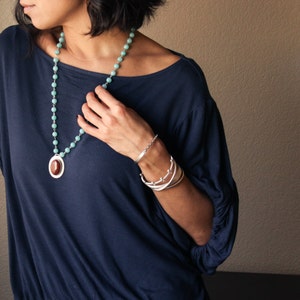 May include: A woman wearing a navy blue top and jewelry. The jewelry includes a long necklace with turquoise beads and a pendant, and multiple silver bracelets. The bracelets have different designs, including a beaded bracelet and a cuff.