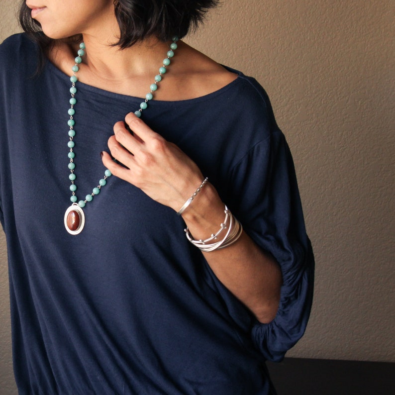 May include: A woman wearing a navy blue top and jewelry. The jewelry includes a long necklace with turquoise beads and a pendant, and multiple silver bracelets. The bracelets have different designs, including a beaded bracelet and a cuff.