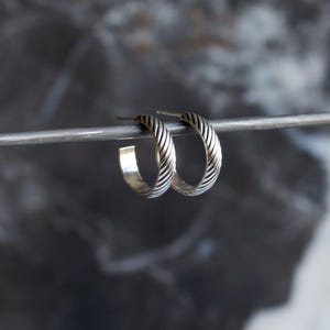 May include: A pair of silver hoop earrings with a textured, ridged design. The earrings are displayed on a silver bar against a blurred, dark background. The hoops have a classic, circular shape and a polished finish.